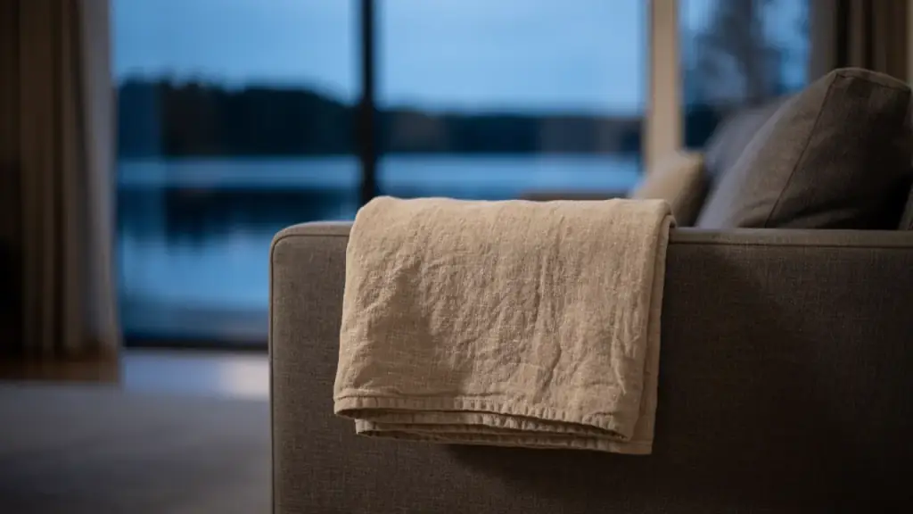 A folded linen throw on the arm of a grey sofa, a lake at dusk out of focus through the windows behind
